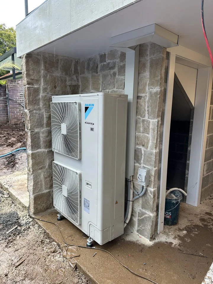 Tall Daikin outdoor air conditioning unit with insulated pipework against a stone wall