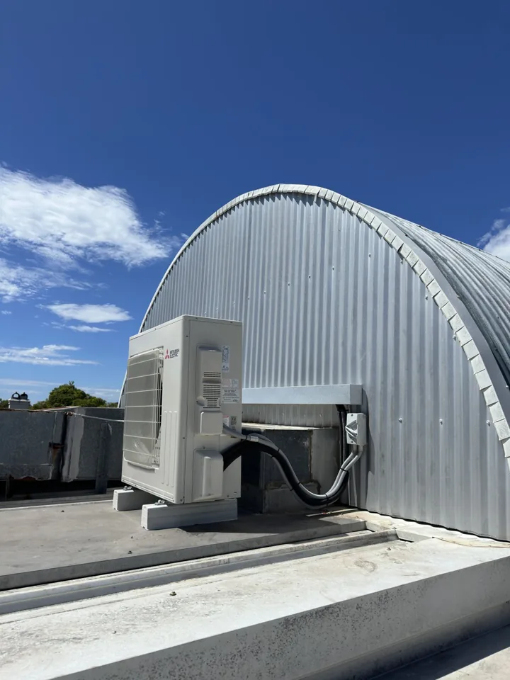 Outdoor air conditioning unit installed on a flat roof beside a curved metal building