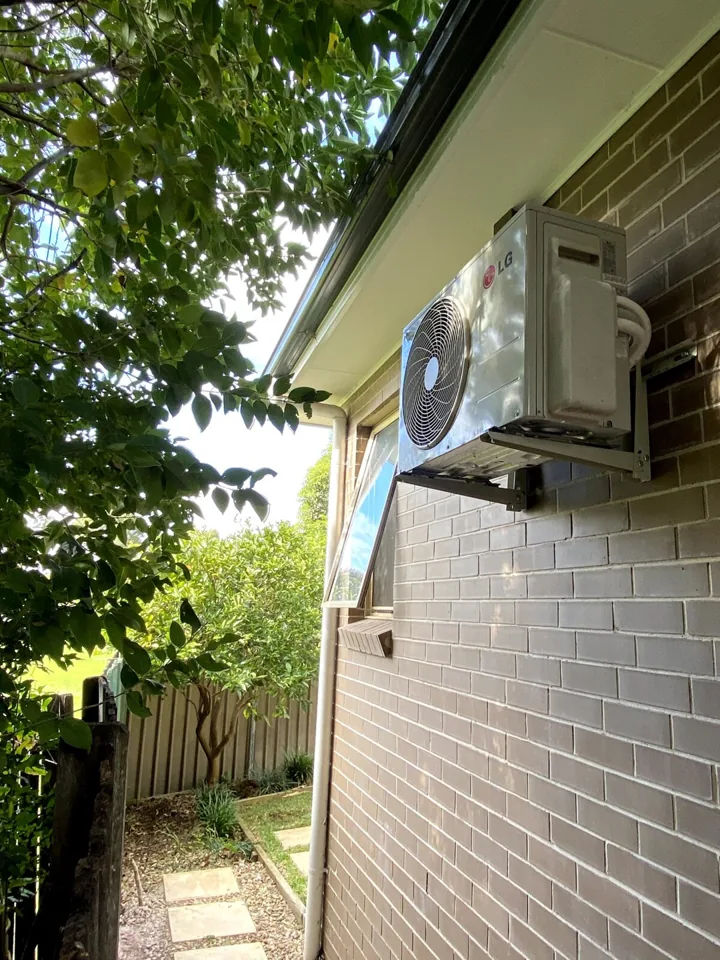 LG outdoor air conditioning unit installed on wall brackets beneath the eaves of a brick home